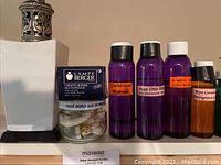 White ceramic Lampe Berger catalytic burner with parts and four purple and small orange bottles of essential oils arranged side by side.