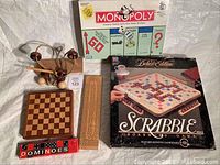 Photo showing various boxed and loose games including Monopoly, Scrabble Deluxe Edition, magnetic chess set pieces in wooden box, dominoes, cribbage board, and wood puzzle toy.