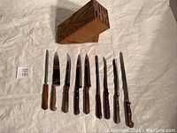 Photo showing 8 kitchen cutting knives arranged beside a wooden knife block on a white cloth background