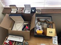 Overview of multiple boxes containing assorted women's jewelry and perfume items on table near window.