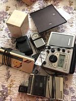 Overview of various radios, cameras, and a multifunctional handheld radio with analog and digital display, along with a compact camera in case and boxed item.