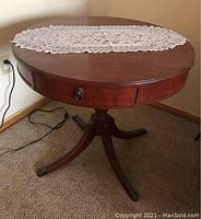Full view of the round Duncan Phyfe style wood pedestal table with a lace runner on top, showing brass hardware and three curved legs.
