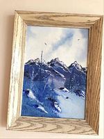 Framed painting of a snowy mountain landscape with blue sky and clouds, natural wood frame.