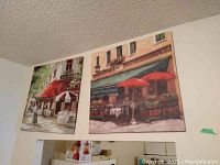 Two 24x24 canvas prints hung above a doorway showing outdoor café scenes, one with white umbrellas and one with red umbrellas, floral window boxes, and chairs.