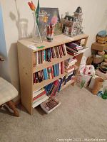 Wide view of books on wooden shelf with books stacked and standing, 3D puzzle of church building on top near vase and cup with pens