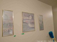 Three framed prints hanging on a wall, arranged side by side. Middle print larger depicting a house, two slimmer prints with waterside scenes on each side.