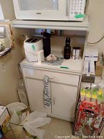 Full view of white MDF microwave cart holding a microwave oven on top, with items on the shelf and towel hanging on the side. The cart has a cabinet with doors.