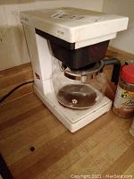 White Mr. Coffee drip coffee maker on kitchen counter with glass carafe and black handle, showing stained and worn condition.