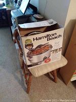 Hamilton Beach slow cooker in its original box sitting on a chair, showing brand and model on front.