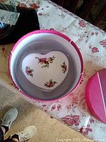 Heart shaped porcelain trinket box with small rose floral pattern inside a pink round box on a floral tablecloth.