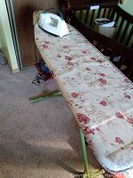 Iron resting on top of the floral plastic-covered ironing board with green legs in a room with carpet and wooden furniture.