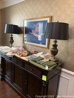 Two table lamps on a dark wood sideboard with dark brown textured drum shades and bronze-finished urn-shaped bases with dark wood square bases, surrounded by fabric and linens.