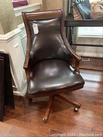 Full view of the leather desk chair showing wooden arms, curved backrest and wooden swivel base with wheels.