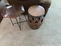 Both side tables placed next to each other on carpet in front of a couch, showing hexagonal metal top table and cylindrical wooden and metal lattice stool.
