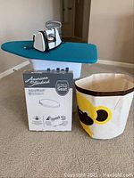 Photo showing small ironing board with teal cover, an iron, white and black carry bag, fabric laundry basket with flower design, and boxed AquaWash bidet seat on carpet.