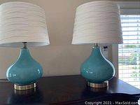 Pair of blue ceramic table lamps with white fabric shades on a dark wooden surface near window with blinds.