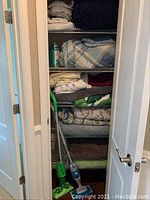 Open closet with multiple folded linens on shelves including blankets, sheets, towels, and two Swiffer floor cleaners at bottom.