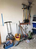 Photo showing garden tools leaning against wall including shovels, rakes, broom, collapsible stair climbing dolly with large wheels, a blue container with miscellaneous hardware and gloves, cordless green and gray EGO string trimmer laying on the floor