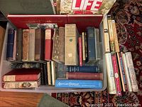 Multiple books arranged in a box and on the floor including worn vintage and antique books with varying colors and sizes.