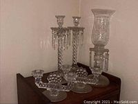 Five glass candle holders on wooden surfaces, showcasing two candelabra holders with hanging crystals, two two-arm geometric pattern holders, and one hurricane etched design holder.