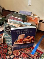 Photo showing an assortment of cookbooks including The Big Book of Breakfast and The Joy of Cooking arranged vertically on a rug.