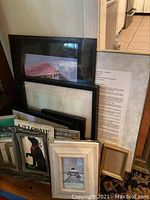 Photo showing several picture frames of different sizes and styles leaning against a wall, including black, white, and silver frames along with some framed documents and photos.