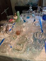 Top-down view showing various glass pitchers, decanters, vases, and bowls on a table with visible blue decoration on one bowl and green frosted decanter.