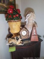 Wide view of entire lot showing clock, dog doorstop, plaster bust, yellow urn with floral arrangement, and metronome.