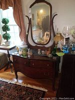 Full front view of wooden vintage vanity with inset marble top, three drawers with brass handles, adjustable oval mirror framed in wood with decorative carving.