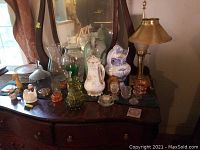 Photo showing five pitchers in various designs, eight stacked dessert plates, antique painted metal duck figurine, three glass flower frogs, Royal Haeger vase with chip, and brass lamp with shade all arranged on a wooden dresser under a mirror.