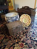 Photo showing wooden magazine rack, ceramic egg holder, wicker chest, and Chinese print planter all placed on patterned carpet.