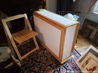 Folded drop leaf table standing upright showing compact folded form with white panels and wood trim on a patterned rug.