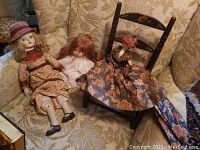 Two porcelain dolls and one wooden doll seated on a floral upholstered chair. Wooden doll missing an arm and wearing floral dress, sitting in a wooden chair with painted flowers.