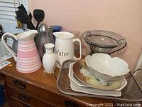 Photo showing ceramic pitchers including pink striped pitcher, white water pitcher, small white vase, metal pitcher with utensils, stainless steel carafe, glass vases, bowls, plate and wire tray arranged on wooden surface.