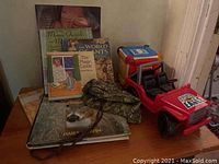 Photo showing a wooden surface with six children's books arranged standing and lying flat, a red toy jeep, a music learning cube, and a camouflage bag.
