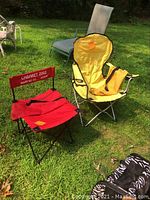 Two folding nylon camp chairs set up on grass outdoors; one small red chair with printed text on backrest and one larger yellow chair with armrests and pockets