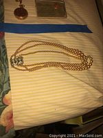 Image of a three-strand faux pearl choker necklace with sterling silver clasp lying on a striped fabric surface. Pearls are cream colored, one strand is visibly missing.