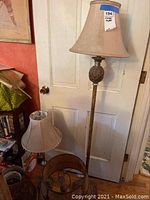 Tall floor lamp with beige fabric shade placed in front of a door, accompanied by a small table lamp with beige shade and a stack of items nearby.