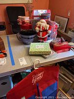 Photo showing a table with two plush Santa figurines, multiple decorative boxes (round and heart-shaped), a large purple bowl, and some ribbons.