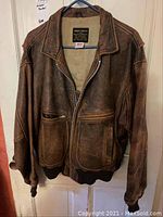 Front view of worn brown leather aviator jacket hung on a door showing front zipper and pockets.
