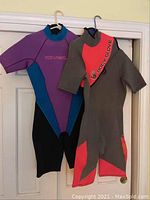 Photo of two short sleeve wetsuits hanging on hangers on a door, showing full length of the suits.