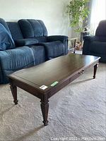 Photo of wooden coffee table in living room setting, showing rectangular top, drawer on one side, and carved tapered legs.