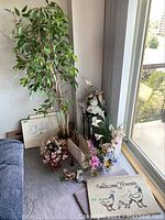 Overview showing artificial tree, flower arrangements, black metal stand, two framed prints, wooden figurine and magazine holder placed near a window in room corner.