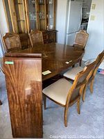 Photo showing wooden dining table with two removable leaves attached, six cane-back chairs surrounding it, light beige cushioned seats, finish damage visible on table top, in room setting.