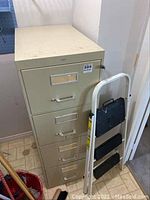 Full view of the metal filing cabinet showing five drawers, white/beige color, stained on top, with the black and white step ladder folded and resting against it.