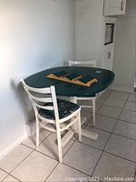 Kitchen table with green top and white pedestal base with two white wooden chairs with patterned cushions placed beside a tiled floor and white wall.