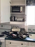 Photo showing small black toaster oven on countertop, matching metal pots and pans with lids, white George Foreman grill, silver two-slice toaster inside cabinet