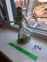 Porcelain figurine of young woman holding bouquet, standing on windowsill with daylight coming through a nearby balcony. Figurine appears in good condition with detailed paint and glazing.