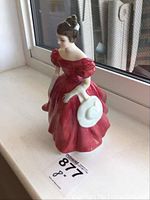 Figurine showing front view of woman in red dress holding white hat on window sill.