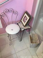 Metal framed chair with white cushioned seat, metal framed stool with floral cushioned top with three framed prints on top, and rectangular waste basket on tile floor against pink wall.